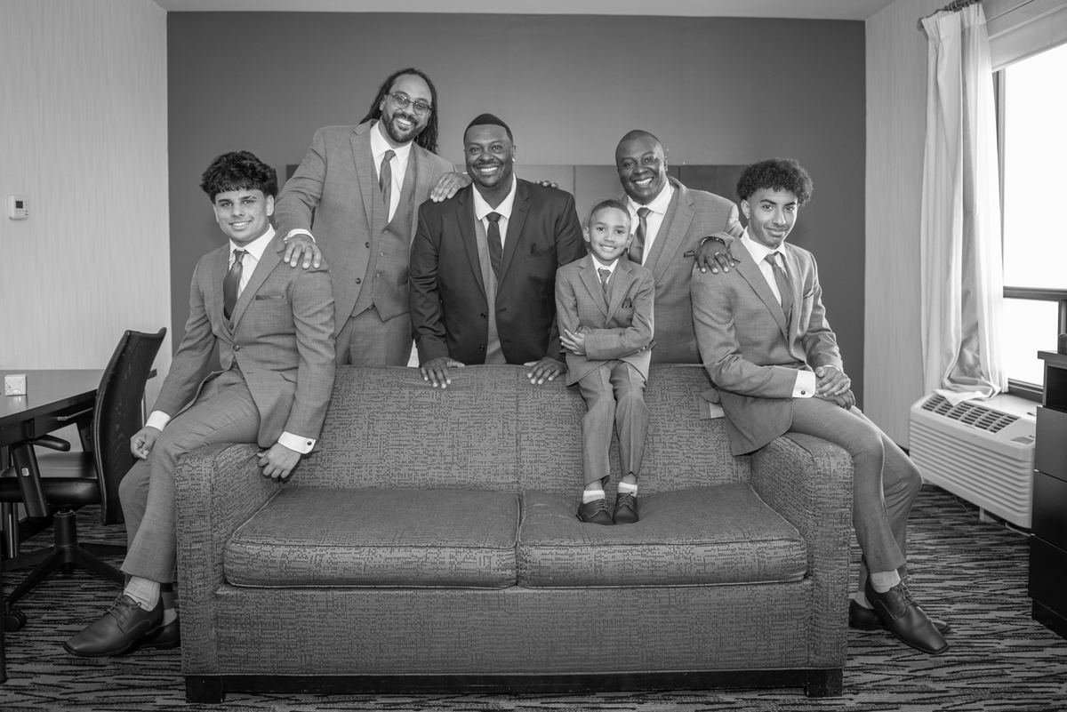 Six men in suits pose on and around a sofa in a hotel room, illuminated by natural light streaming from a window.