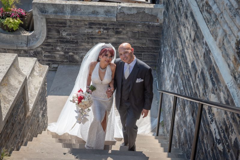 A bride in a white gown and veil with red and white flowers descends stone stairs with her groom in a dark suit.