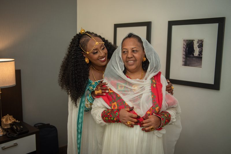Two women in traditional attire embrace warmly under soft indoor lighting, their joyful faces illuminated by a lamp.