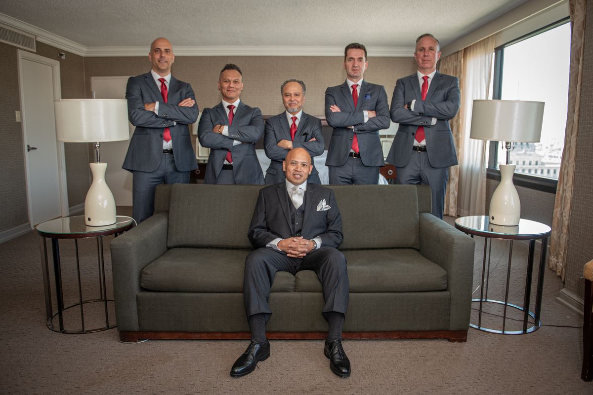 Six formally dressed men pose in a well-lit room, one sitting on a sofa with five standing behind him.