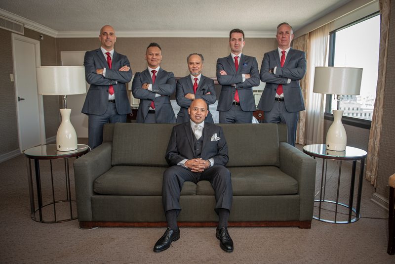 Six formally dressed men pose in a well-lit room, one sitting on a sofa with five standing behind him.