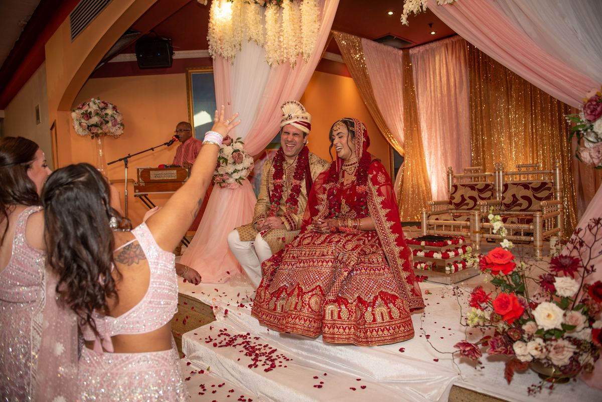 Bride and groom laughing as guests throw rose petals on a decorated stage, surrounded by festive attendees.