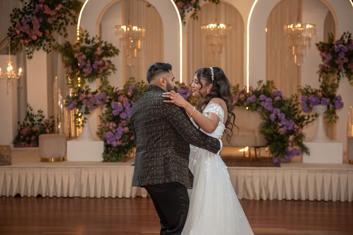 Bride and groom dance intimately on a polished wooden floor, surrounded by floral arches and glowing chandeliers.