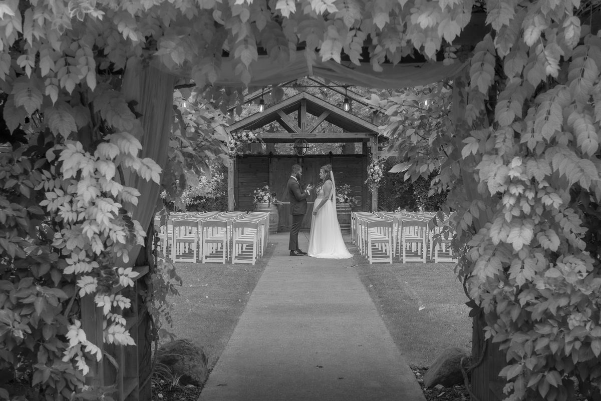 Bride and groom exchange vows under a leafy archway adorned with vines, surrounded by wooden chairs on a paved aisle.