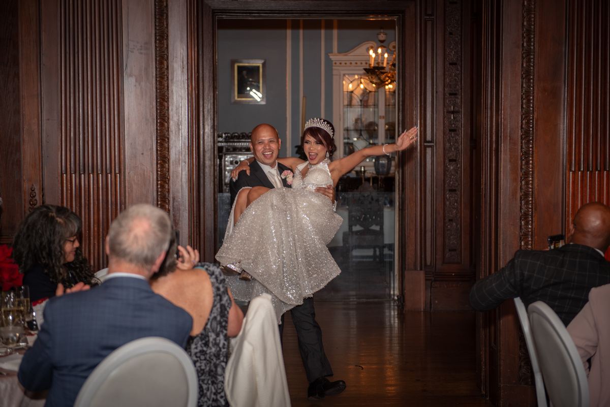 A groom carries his bride through an ornate doorway into a banquet hall illuminated by chandeliers and rich woodwork.
