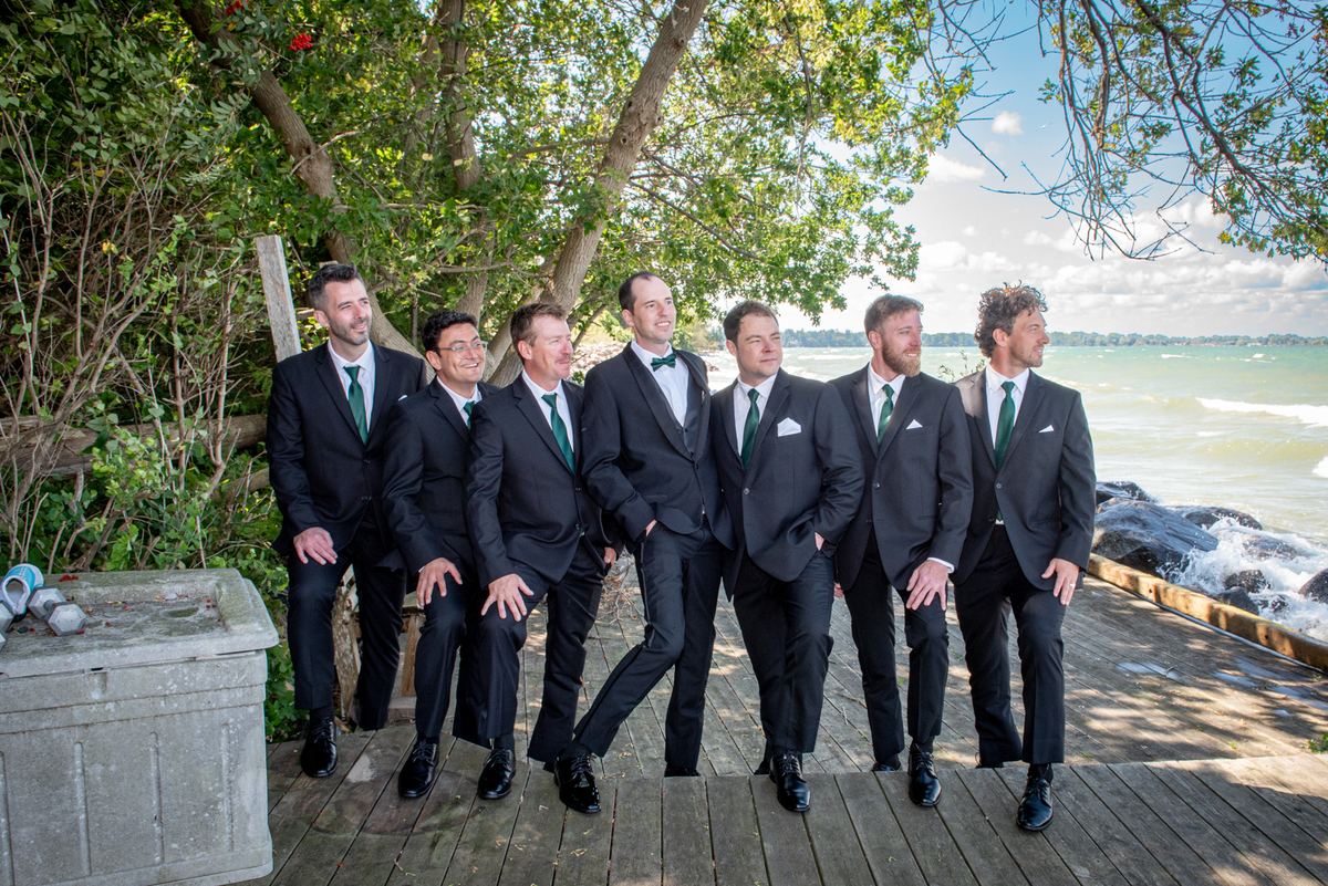 Seven men in black suits and green ties stand smiling on a wooden boardwalk by the sea with crashing waves and trees.