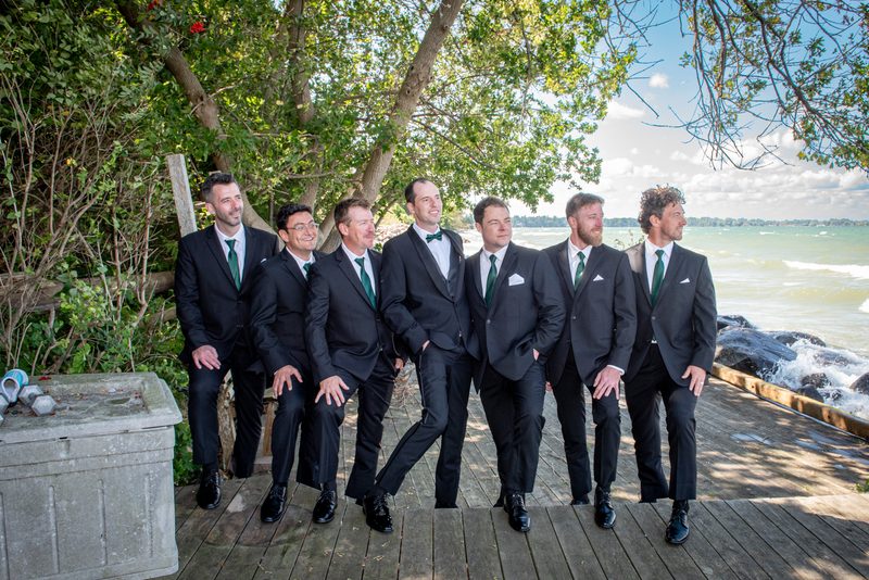 Seven men in black suits and green ties stand smiling on a wooden boardwalk by the sea with crashing waves and trees.