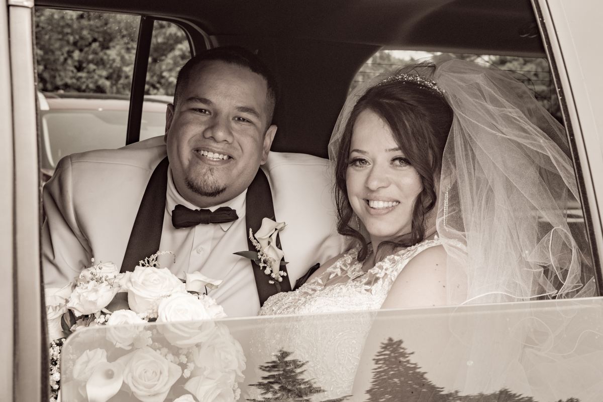 A smiling bride and groom in the backseat of a vintage car, with trees reflected in the rear window.