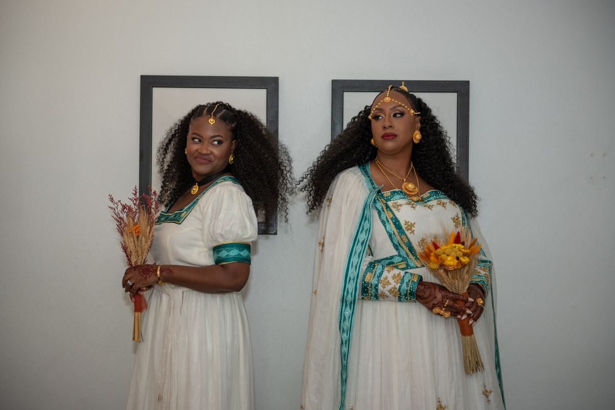 Two women in white and teal traditional outfits hold bouquets and gold jewelry against a plain wall.