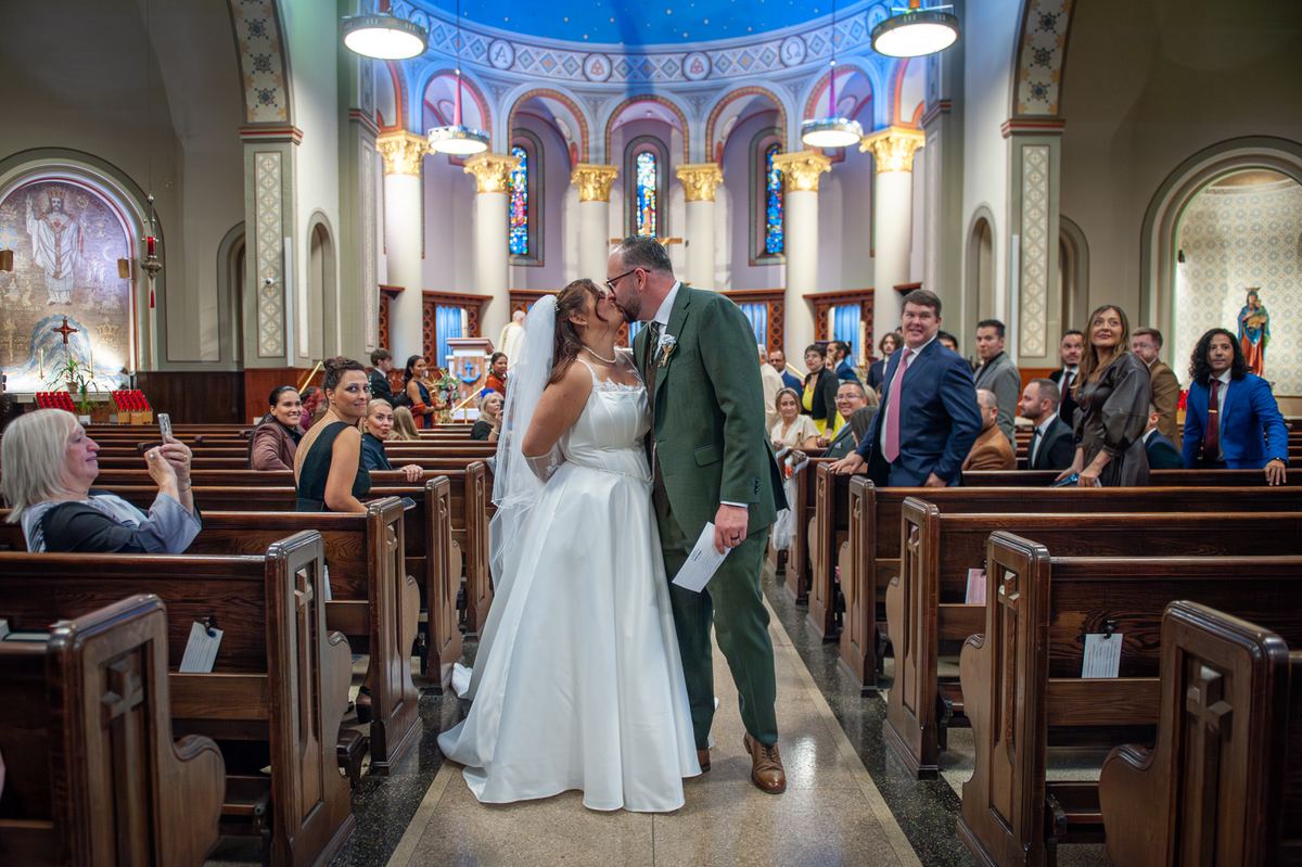 Bride and groom kiss down the aisle surrounded by guests in wooden pews, illuminated by warm lighting from ceiling fixtures.