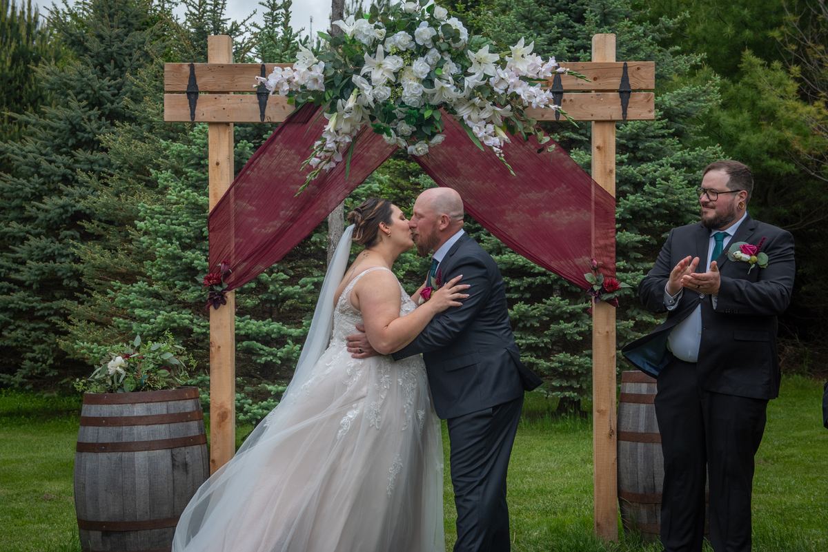 Bride and groom kiss under wooden arch decorated with white flowers and burgundy fabric, framed by evergreen trees.