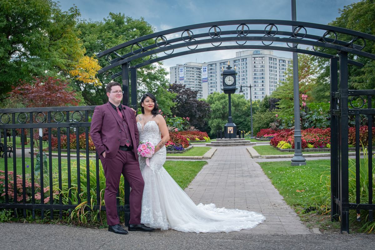 A bride and groom pose under an ornate black metal archway in a park, surrounded by manicured gardens and modern buildings.