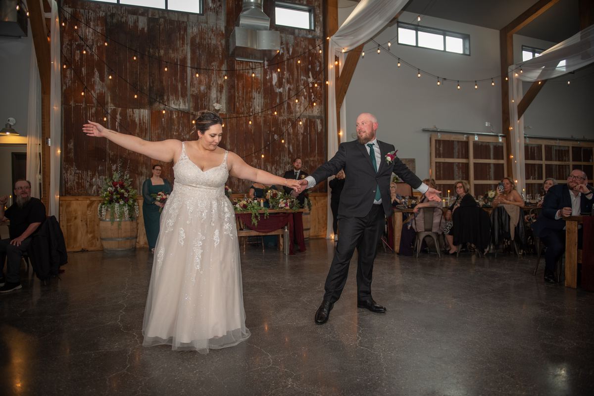 Bride and groom dance joyfully in a rustic setting with warm string lights and smiling guests watching.