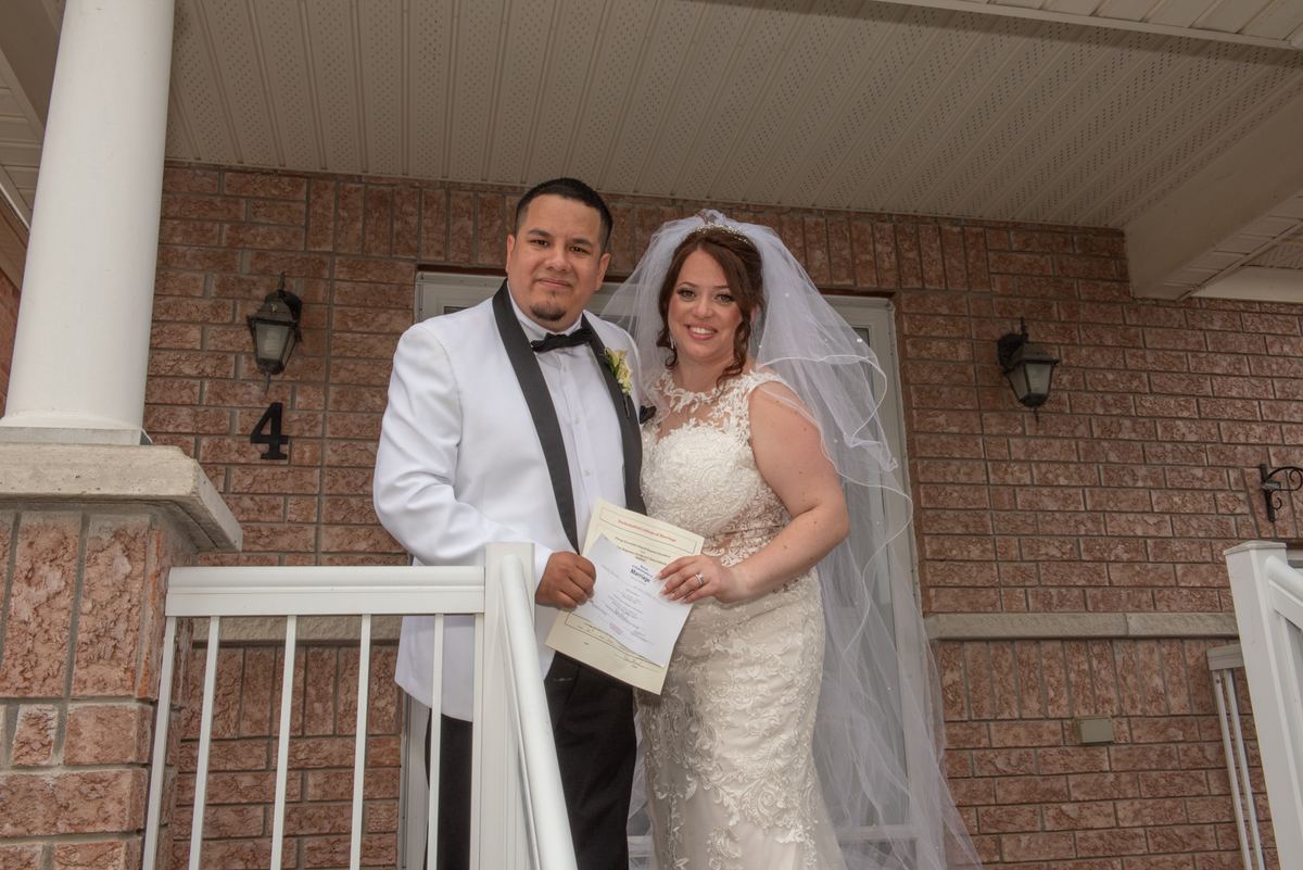 A smiling bride and groom hold their marriage certificate on a porch, framed by a brick wall and white railing.