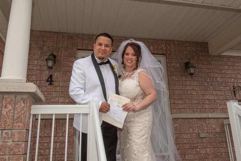 A smiling bride and groom hold their marriage certificate on a porch, framed by a brick wall and white railing.
