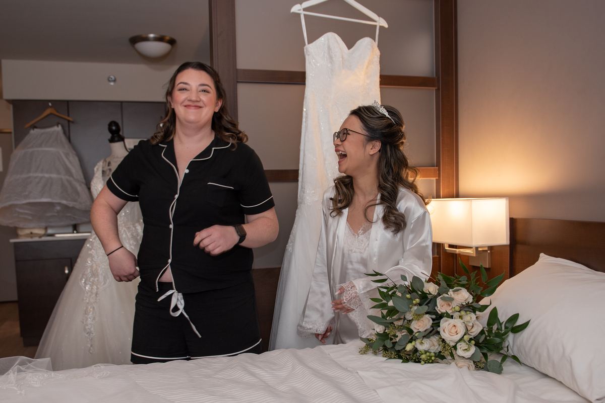 Two women in a bridal suite laugh together by a hanging wedding gown, with a bouquet and additional gowns nearby.