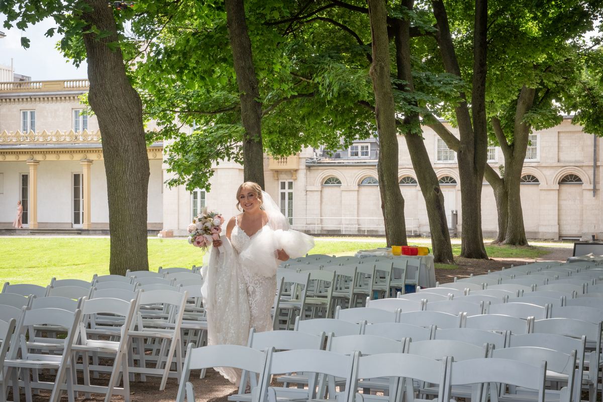 A smiling bride in a courtyard surrounded by white chairs and trees, with sunlight filtering through leaves.