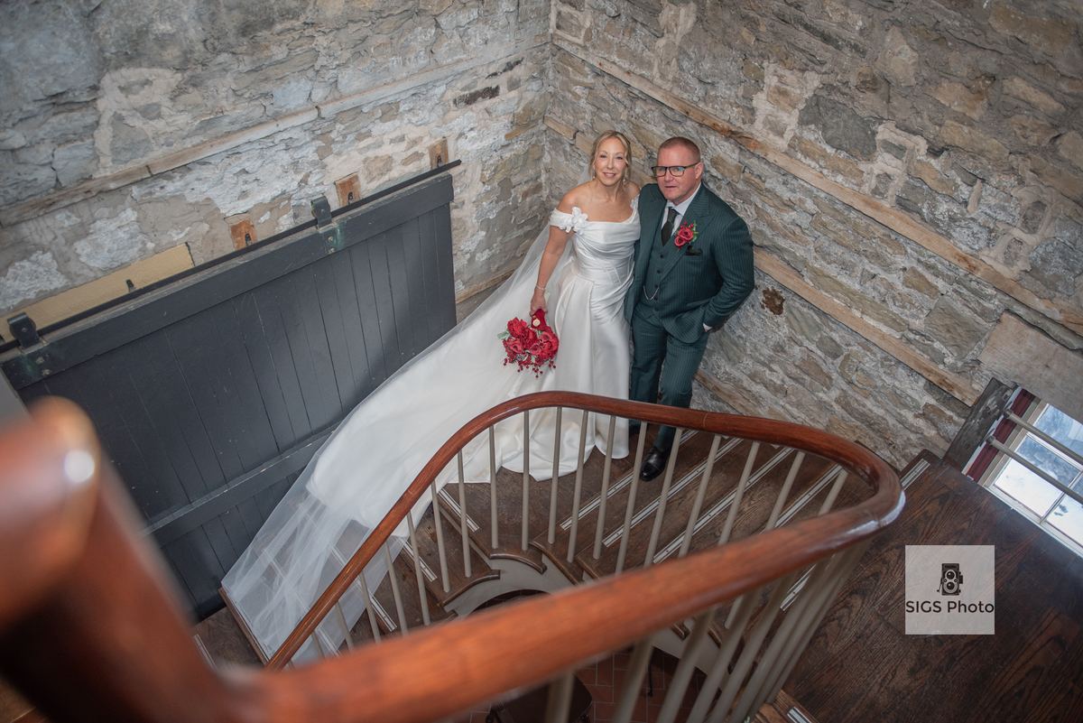A bride in a white gown and groom in a dark suit stand on a spiral staircase inside a stone-walled building.