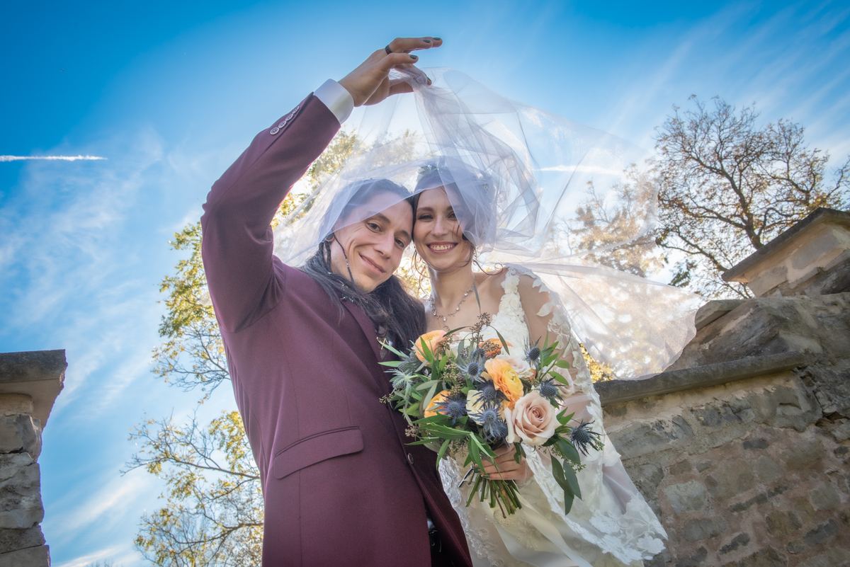 Bride and groom smile warmly outdoors, with the groom lifting her veil as it billows in the wind under autumn trees.