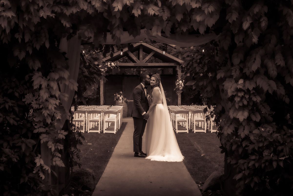A newlywed couple embraces under a leafy archway surrounded by dense foliage and empty chairs leading to a wooden arbor.