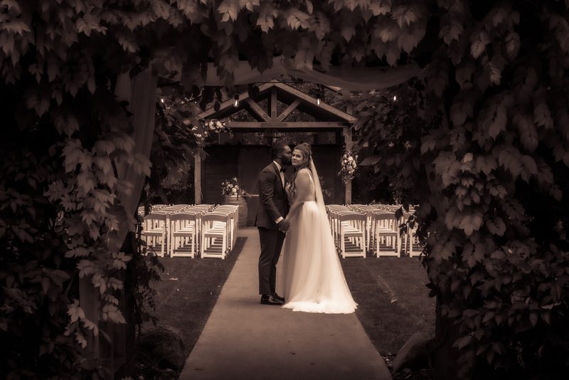 A newlywed couple embraces under a leafy archway surrounded by dense foliage and empty chairs leading to a wooden arbor.
