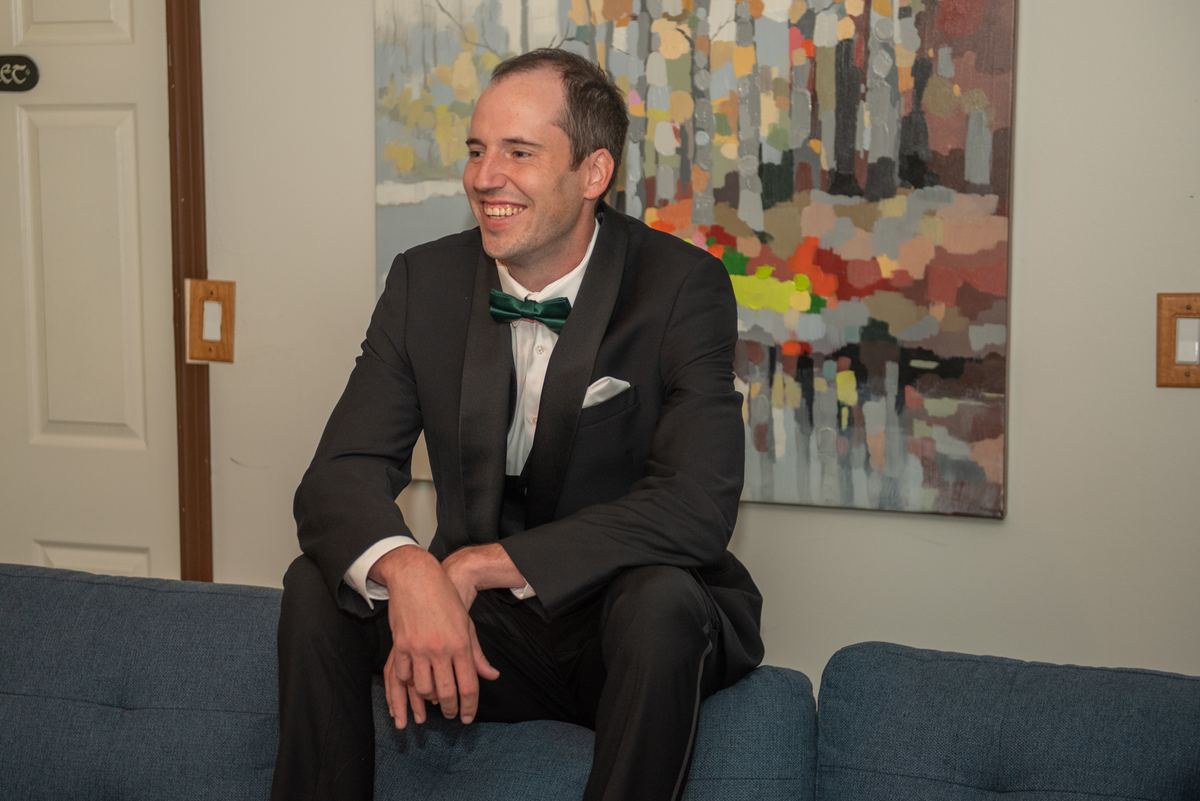 A man in a black tuxedo with a green bow tie sits smiling on a blue couch, next to an abstract painting and a white door.