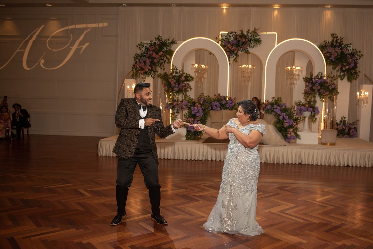 A joyful couple dances on a wooden floor, surrounded by an opulent wedding backdrop with arched lighting.