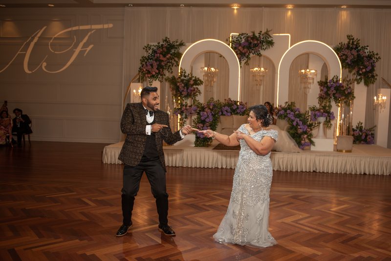 A joyful couple dances on a wooden floor, surrounded by an opulent wedding backdrop with arched lighting.