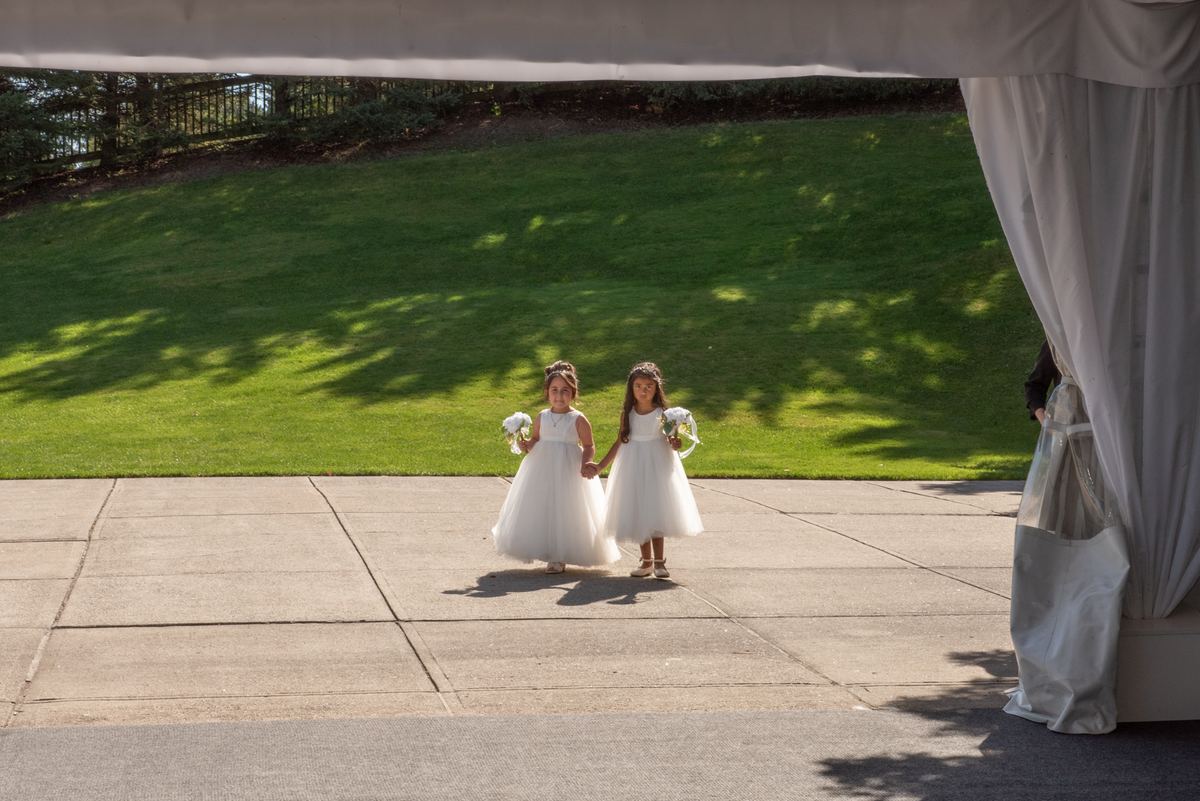 Two young flower girls in white dresses walk hand-in-hand under a draped canopy, each holding small bouquets.