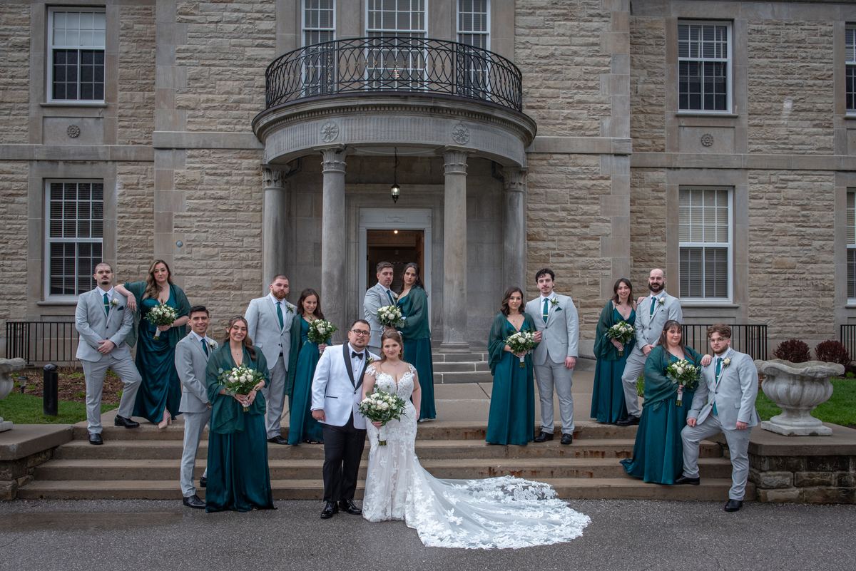 Emily and Ramon stand together on the stone steps of an elegant building, surrounded by their wedding party.