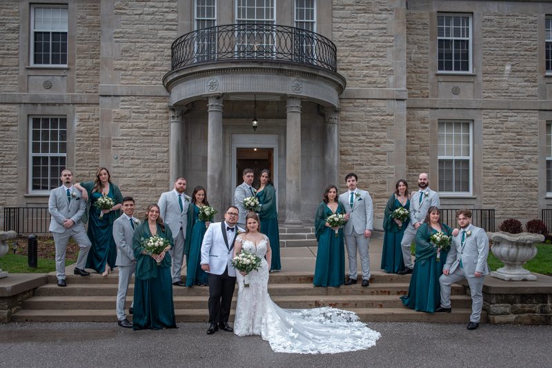 Emily and Ramon stand together on the stone steps of an elegant building, surrounded by their wedding party.