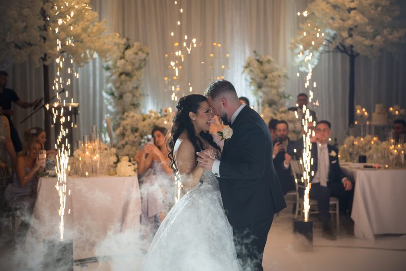 A bride and groom dance amidst swirling fog and sparklers, with guests watching from decorated tables.