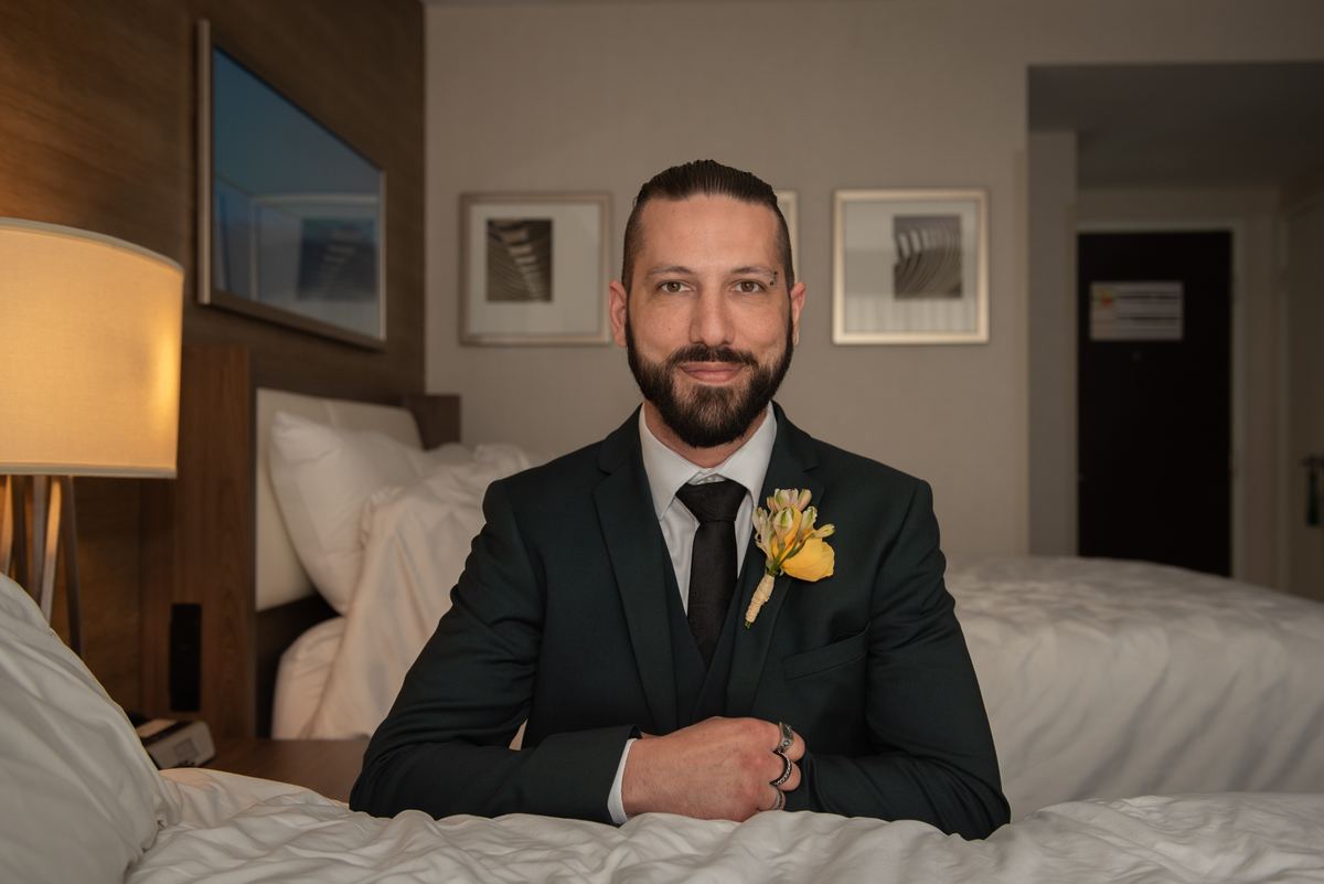 A groom in a dark suit smiles at the camera on a hotel bed, bathed in warm light from a bedside lamp.