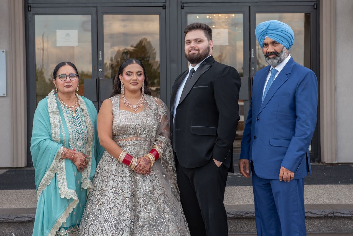 A formal family portrait outdoors features a bride in an ornate silver lehenga, groom in black tuxedo.