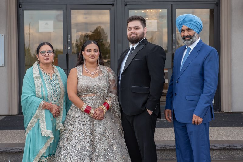 A formal family portrait outdoors features a bride in an ornate silver lehenga, groom in black tuxedo.