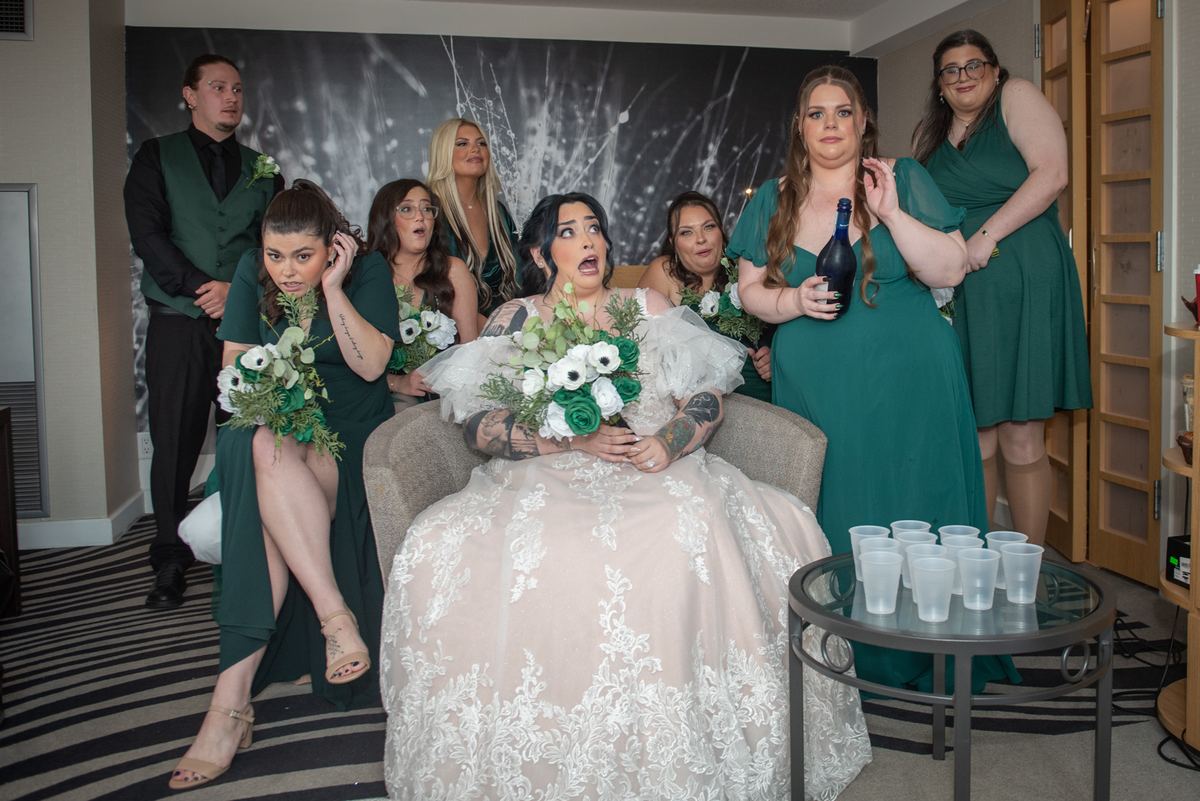 A bride sits with her surprised wedding party, including bridesmaids in green dresses and a groom in a green vest.