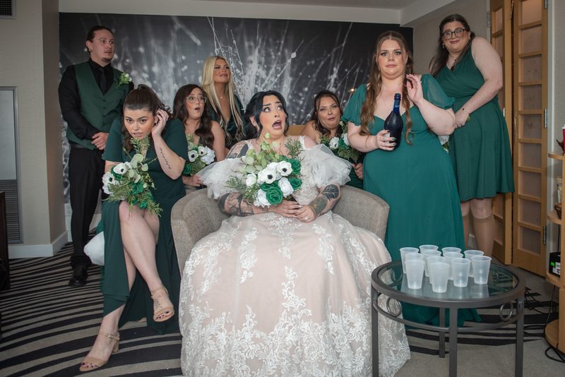 A bride sits with her surprised wedding party, including bridesmaids in green dresses and a groom in a green vest.