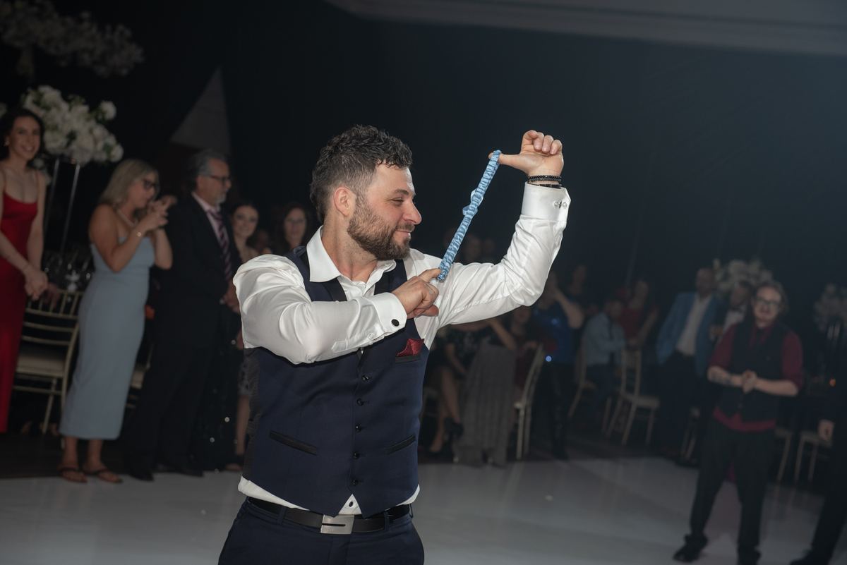 A man in formal wear twirls a blue ribbon joyfully on stage during a celebration, guests clapping and smiling.