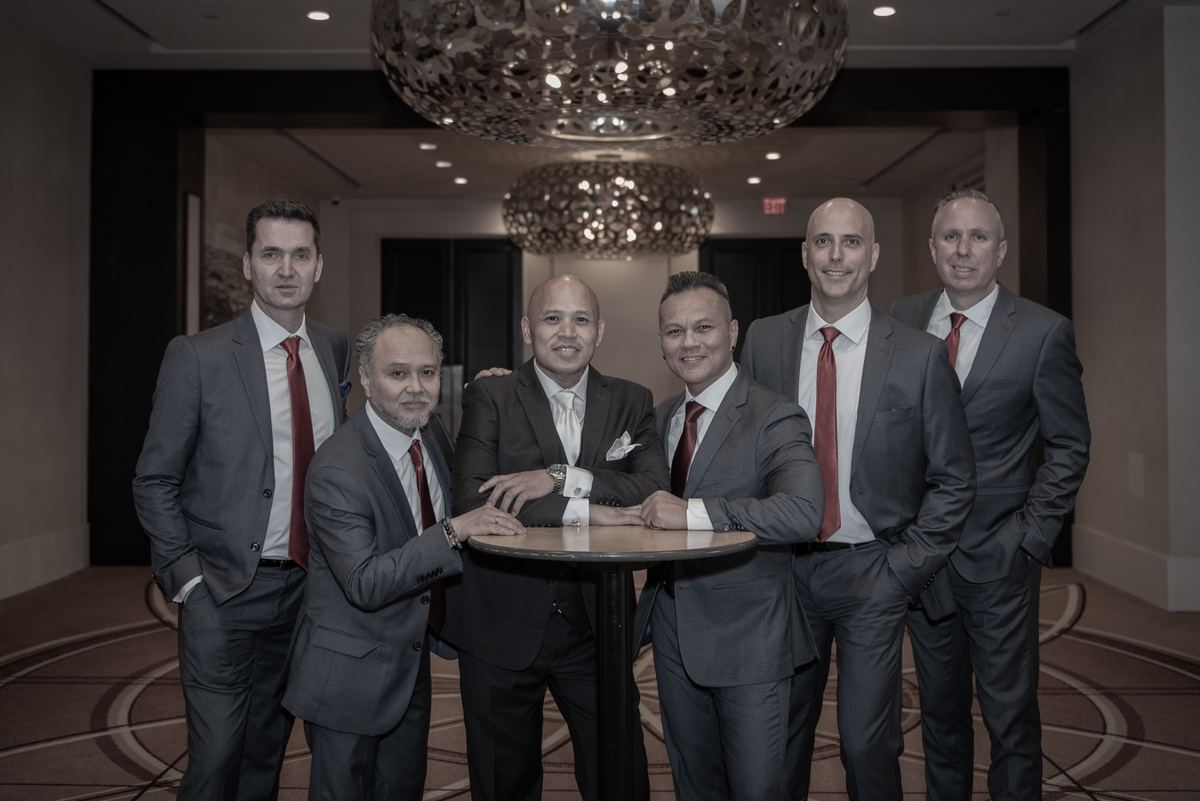 Six men in suits and red ties pose around a table in a luxurious hotel lobby, illuminated by ornate chandeliers.