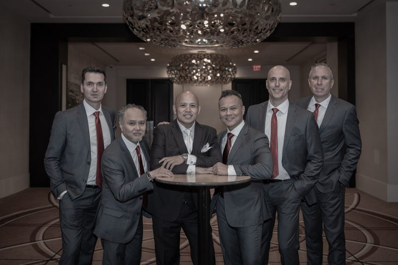 Six men in suits and red ties pose around a table in a luxurious hotel lobby, illuminated by ornate chandeliers.