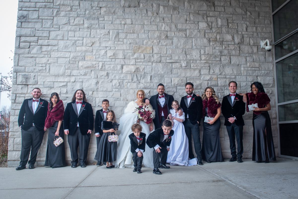 A formal wedding group stands outdoors against a stone wall in elegant attire with burgundy accents.