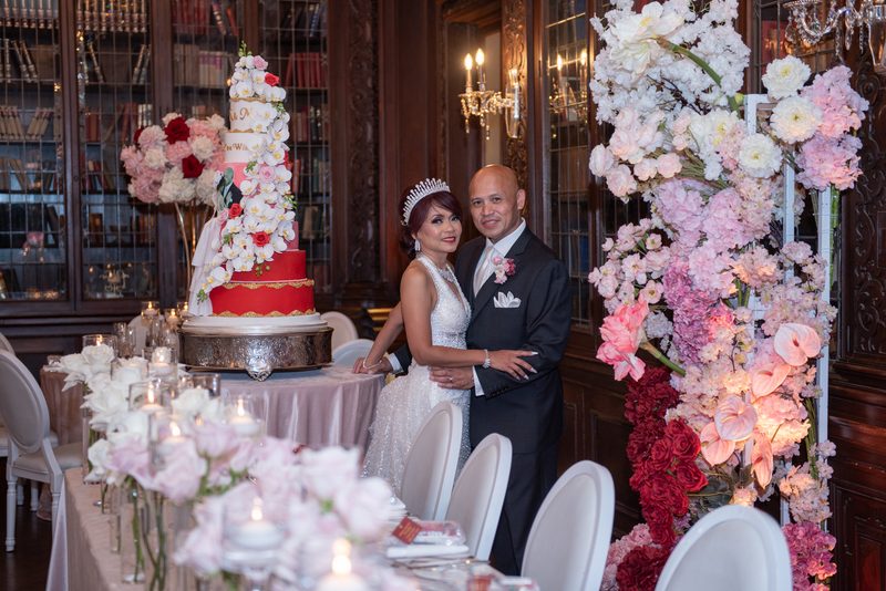 A smiling bride in a white gown and tiara stands with her tuxedoed groom beside a multi-tiered cake and floral arch.