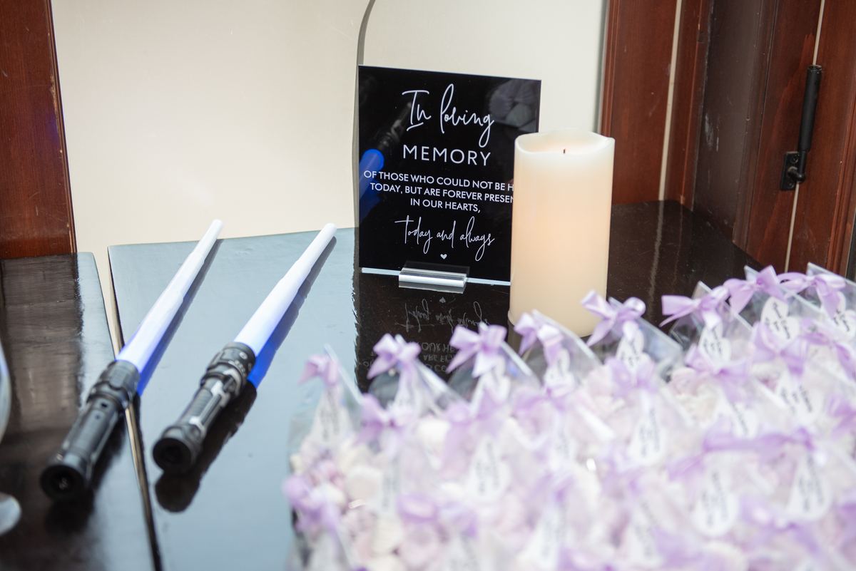 A dark reflective surface holds two lightsabers, a lit candle, and a memory sign under soft lighting.
