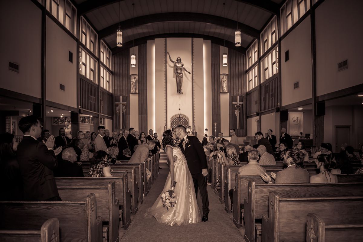 A newlywed couple kisses at the altar, surrounded by applauding guests in sepia-toned light from tall windows and fixtures.