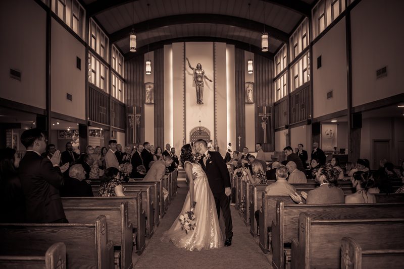 A newlywed couple kisses at the altar, surrounded by applauding guests in sepia-toned light from tall windows and fixtures.