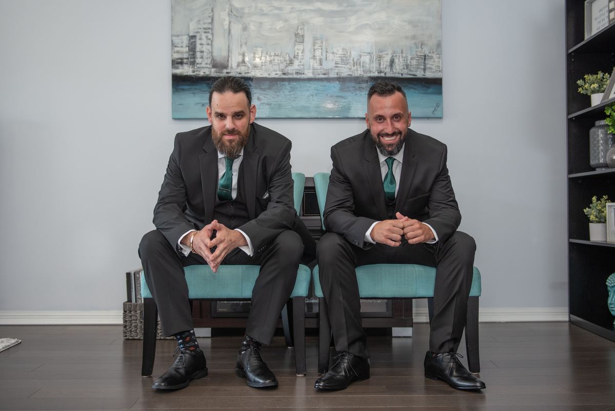 Two men in dark suits and teal ties smile at the camera in a modern room with an abstract cityscape painting behind them.