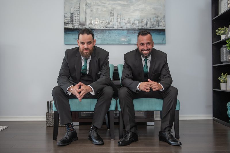 Two men in dark suits and teal ties smile at the camera in a modern room with an abstract cityscape painting behind them.