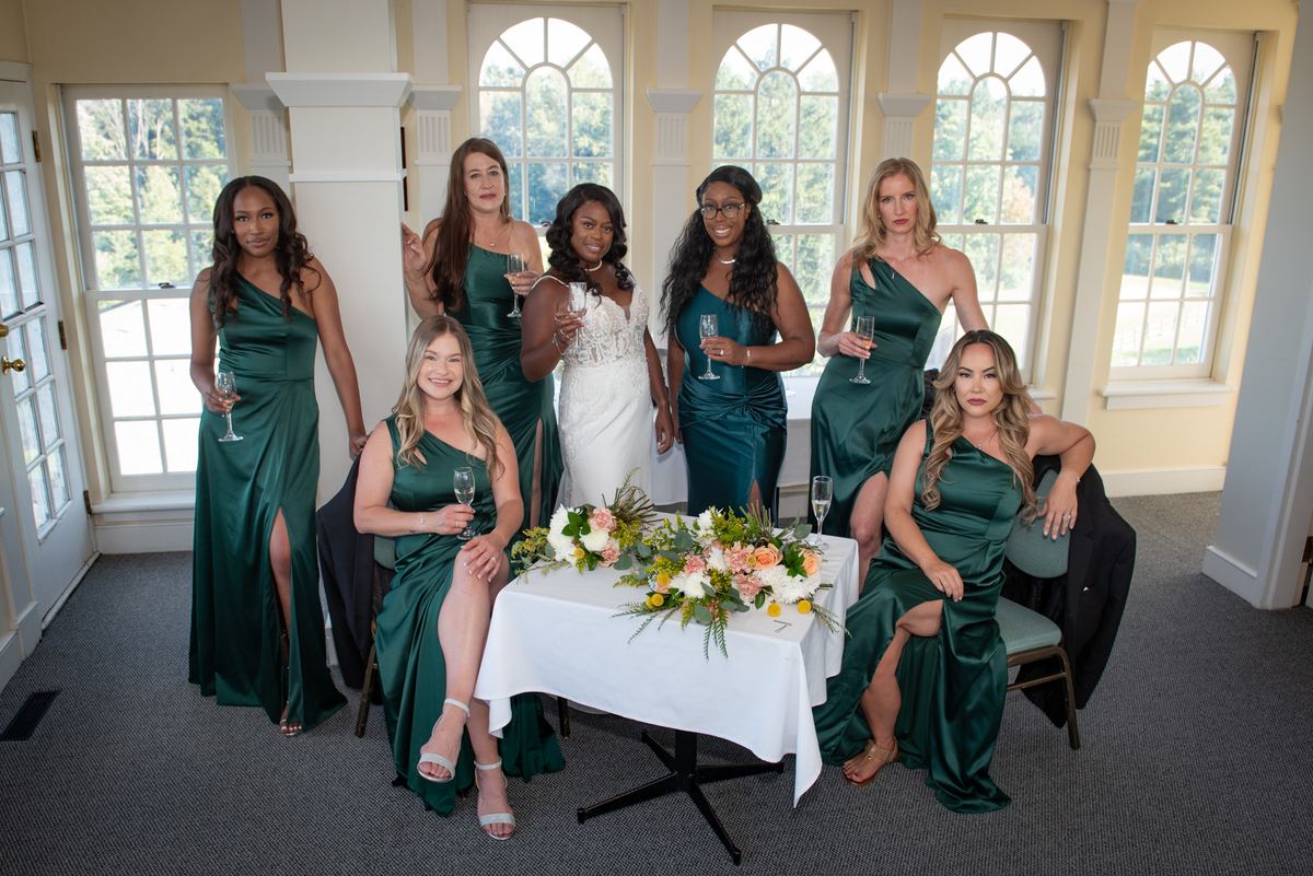 A bride and her bridesmaids in emerald green dresses hold champagne flutes around a table with flowers.