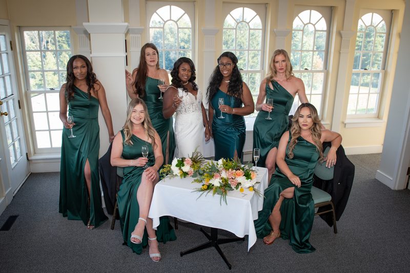 A bride and her bridesmaids in emerald green dresses hold champagne flutes around a table with flowers.