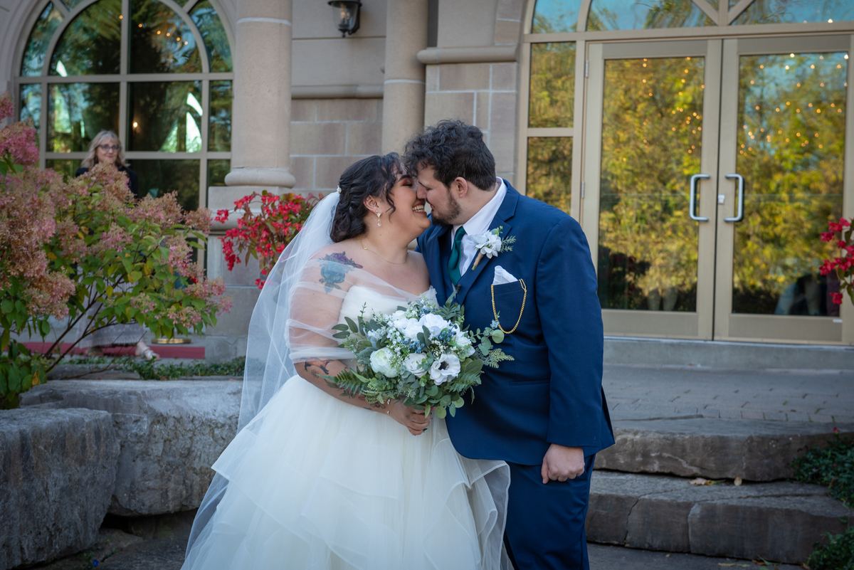 A bride in a white gown and veil kisses her groom in front of stone steps, autumn foliage, and festive string lights.